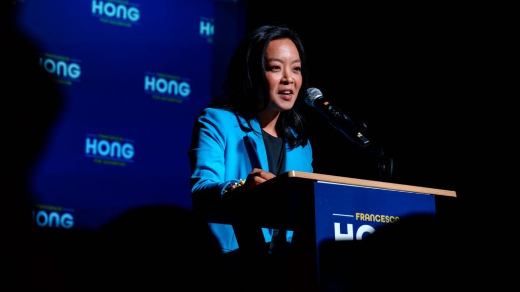 Image of Francesca Hong, a Wisconsin state assemblywoman and Democratic candidate for governor, facing right and speaking to a crowd behind a lectern. She's wearing a pale blue jacket and dark blue shirt