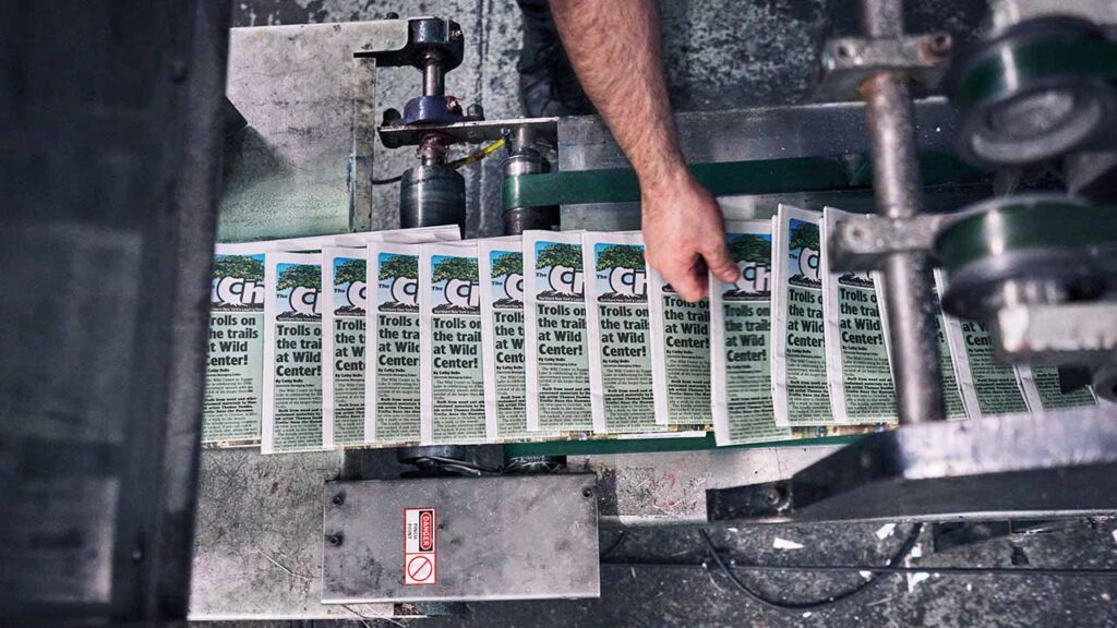 Copies of The Chronicle as they come off the press at the Sun Community News and Printing in Elizabethtown, N.Y., on July 8, 2026. The paper’s advertisers are local businesses, and each week, their ads are distributed in 20,000 dead-tree copies and a digital replica version that gets blasted out to 20,000 email recipients. (An Rong Xu/The New York Times)