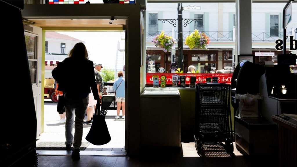 A visitor walks out with a bag of groceries from Doud's Market in Mackinac Island, Mich., June 8, 2026. In the nearly two years since President Trump clinched his return to office, the U.S. economy has come under great duress, leaving families and businesses to suffer through the expensive consequences of constant global conflict and soaring government debt. (Emily Elconin/The New York Times)