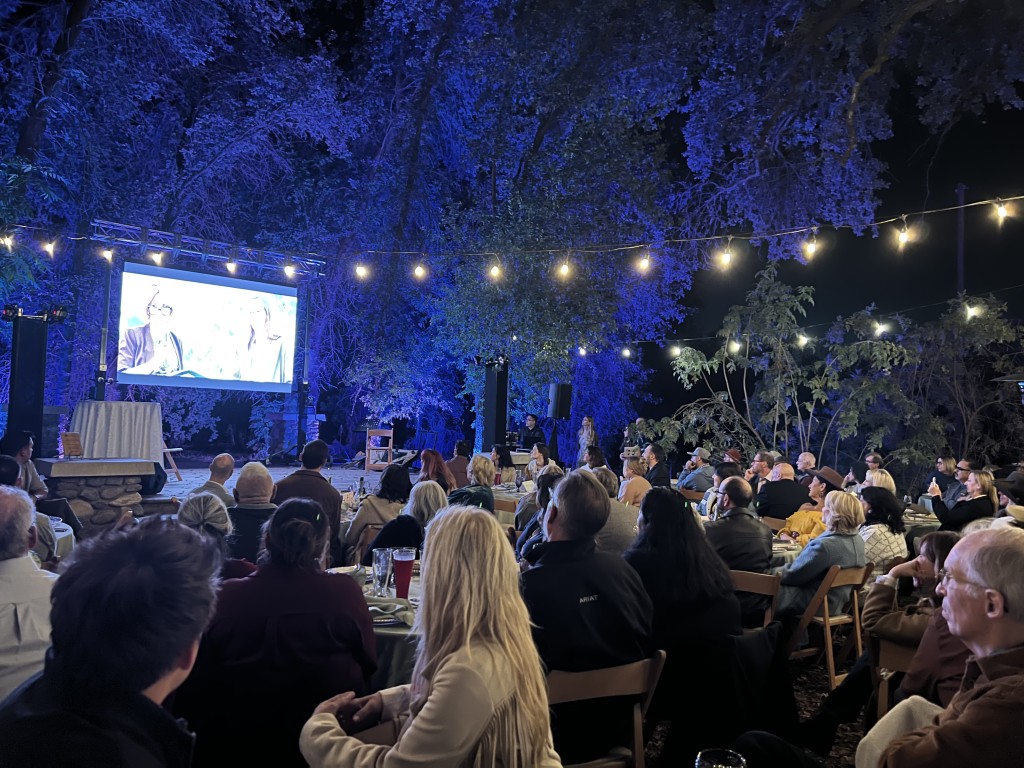Image of a gala under the stars cast in a blue hue with people seated and looking at a video board 