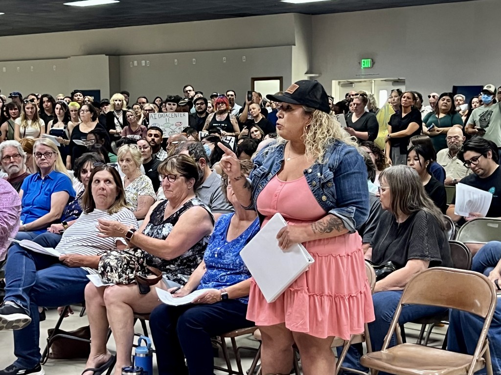 Image of data center protesters packed into a hearing room with a woman in the front row, wearing a pink dress, standing and making a point with her finger
