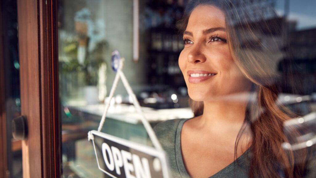 Image of a young woman facing left and looking through the glass of a business with an "open" sign
