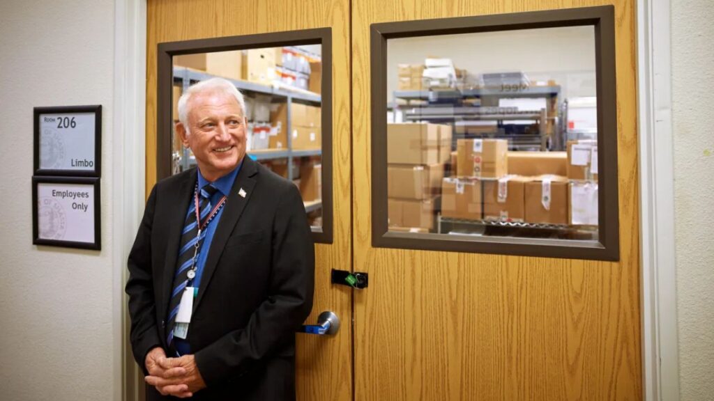 Image of a smiling man in a dark suit looking right and smiling as he leans against two locked maple-colored doors with windows