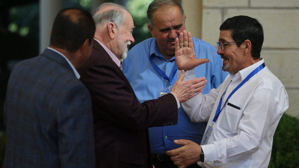 Image of a man in a burgundy suit facing right and greeting a man in a white shirt as a man in a blue shirt looks on