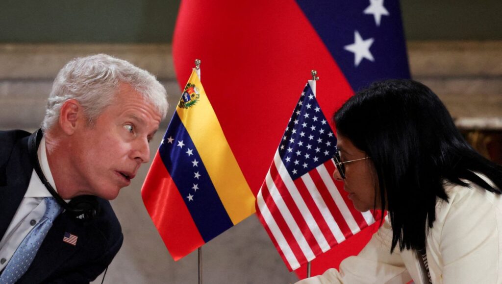 Image of a man in a blue suit with a US flag lapel pin facing right and talking to a woman in a white suit against a backdrop of the Venezuelan and US flags