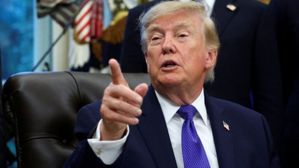Image of President Trump in a dark suit, white shirt, and purple tie, speaking and pointing to a questioner with his index finger