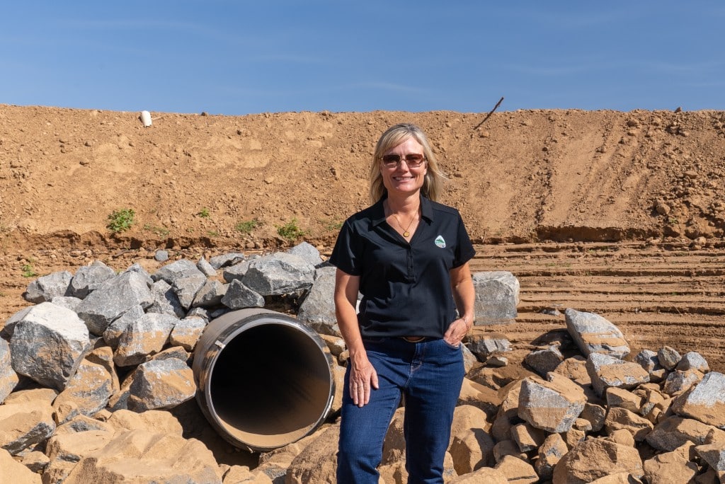 Image of a woman in a blue shirt and jeans standing in an excavated basin near a large water pipe and a rockpile