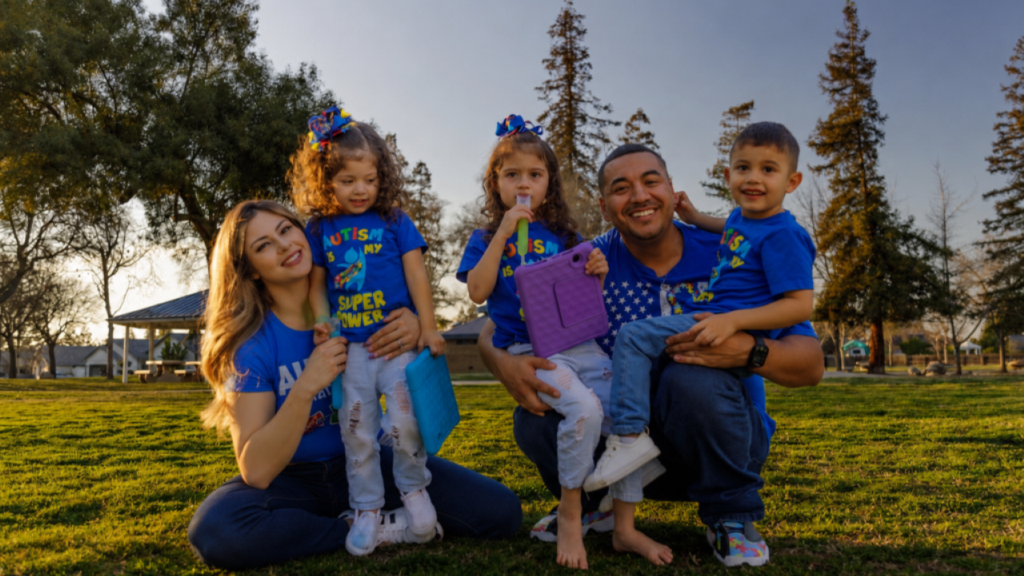 Image of a husband and wife holding three children and posing for a picture on a green lawn in a park