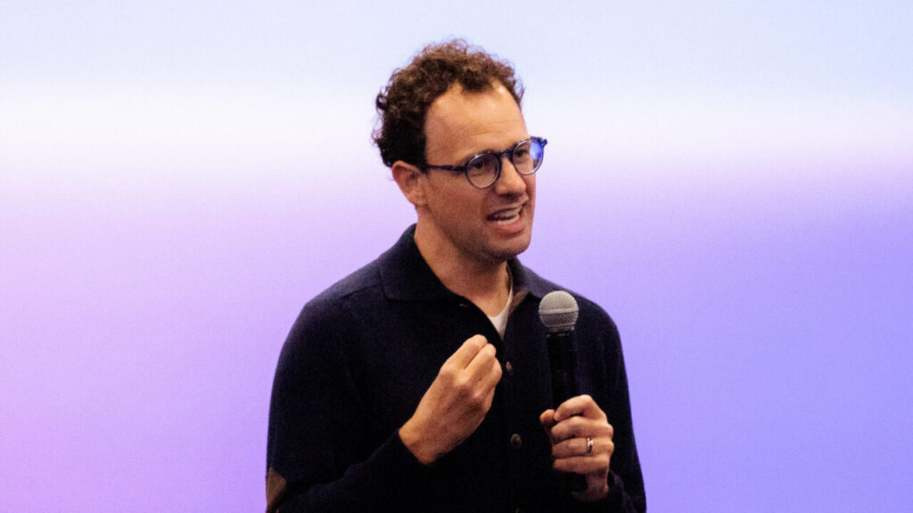 Image of a man in a black shirt facing right with a microphone in one hand speaking to a crowd against a purple, blue, and white backdrop