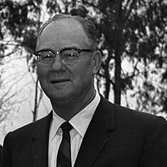 Black and white image of the American developer Del Webb in a dark suit and tie with a white shirt. He is facing left and wearing glasses.