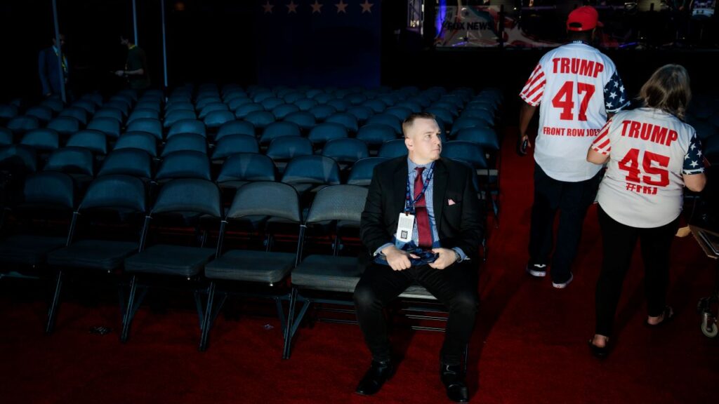Image of a man in a blue suit sitting and facing the camera, along with two people, their backs to the camera, wearing pro-Trump t-shirts
