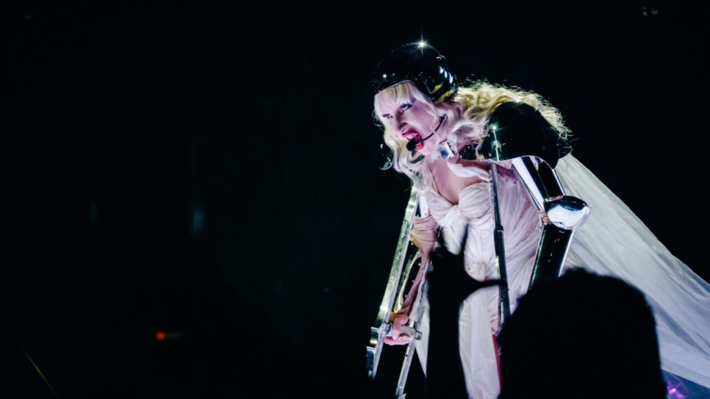 Image of Lady Gaga in white with a black helmet facing left and bending down while singing against a black backdrop