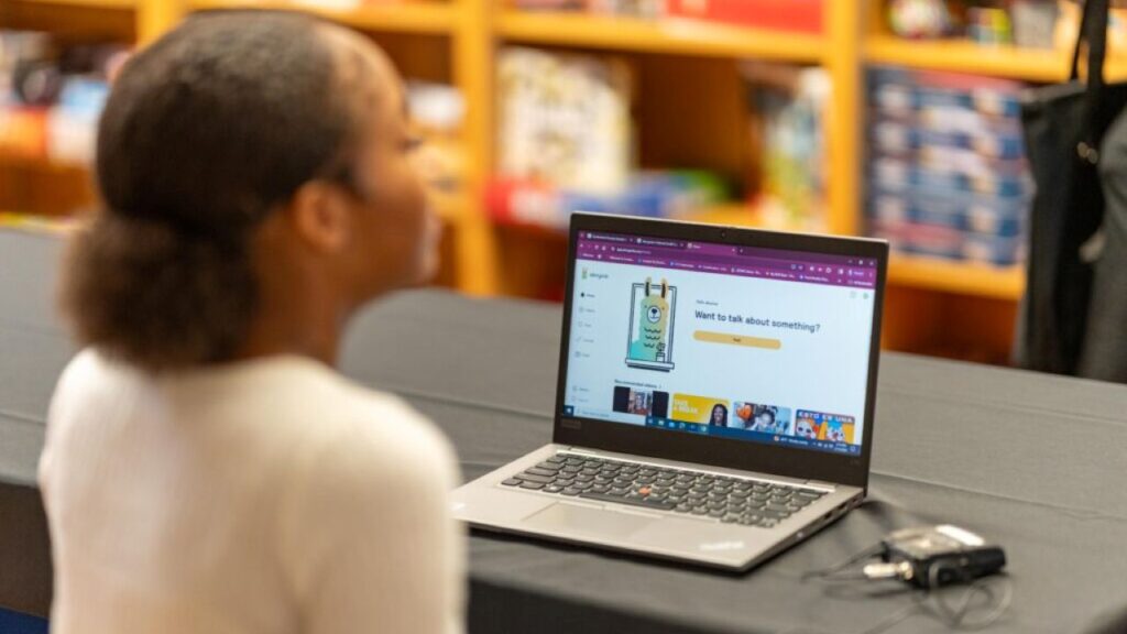 Image of a female student in a light colored sweater facing right and engaging with a chatbot via a notebook computer