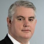 Portrait of Sean Higgins in a dark coat, white shirt, and red tie facing straight ahead
