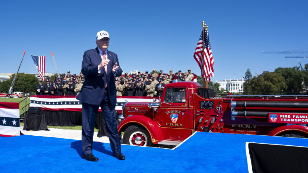 Image of President Trump in a USA truckers hat and blue suit standing on a stage near a red firetruck and clapping his hands