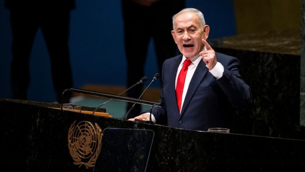 Image of Benjamin Netanyahu in a blue suit, white shirt, and red tie, pointing as he speaks