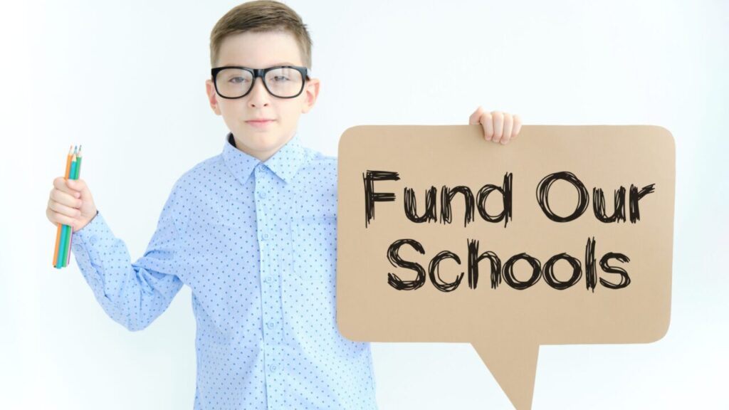 Image of a young student in a blue shirt and oversized black-rimmed eyeglasses holding a sign stating "Fund Our Schools"