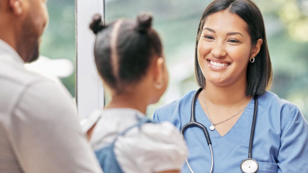 Image of a smiling female nurse in blue scrubs with a young patient and her father, symbolizing high-quality healthcare