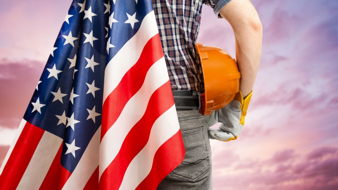Image of the back of a worker who is holding an American flag and an orange hard hat, symbolizing Labor Day