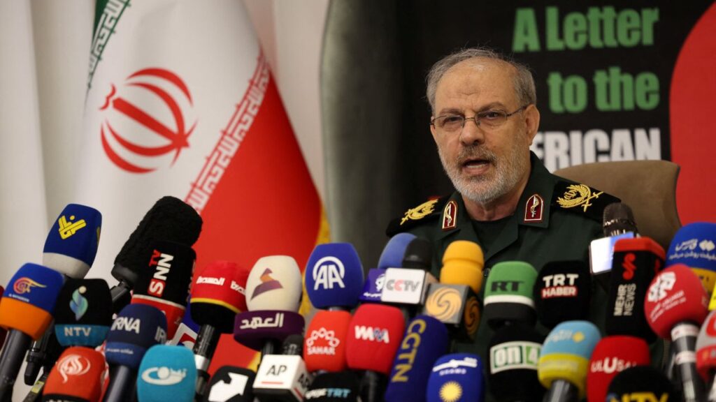 Image of IRGC spokesperson Hossein Mohebb speaking behind a desk covered in media microphones