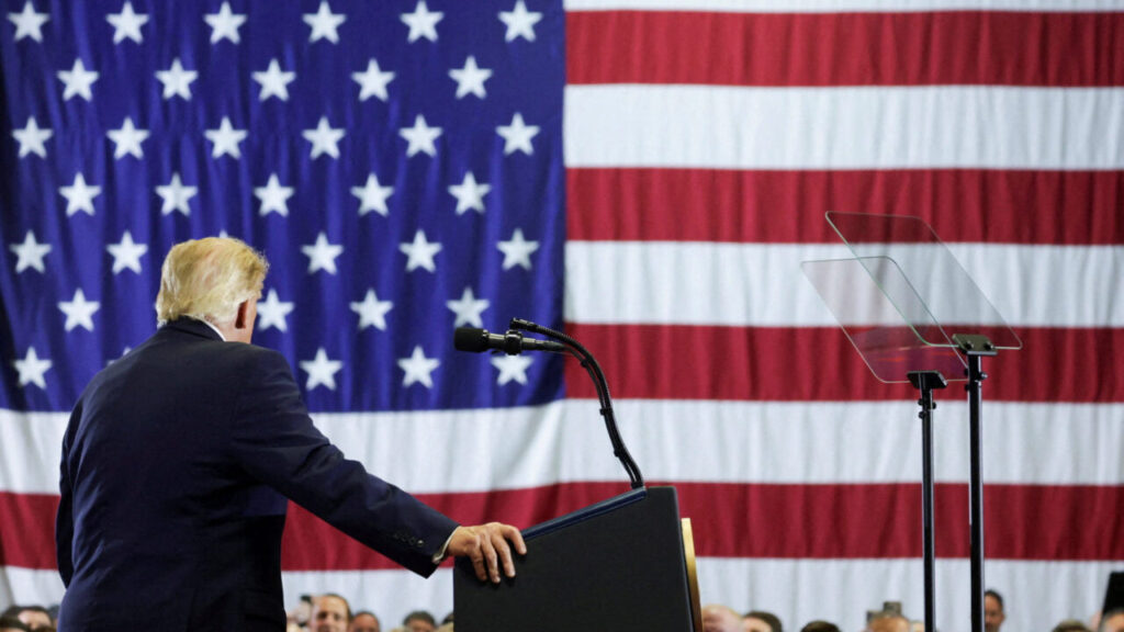 Image of President Trump in a dark suit with his back to the camera, leaning on a podium and talking to a crowd against an American flag backdrop