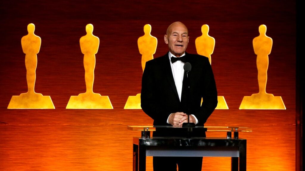 Image of Patrick Stewart standing on stage in a tuxedo and speaking against an orange-hued backdrop of Academy Awards statues