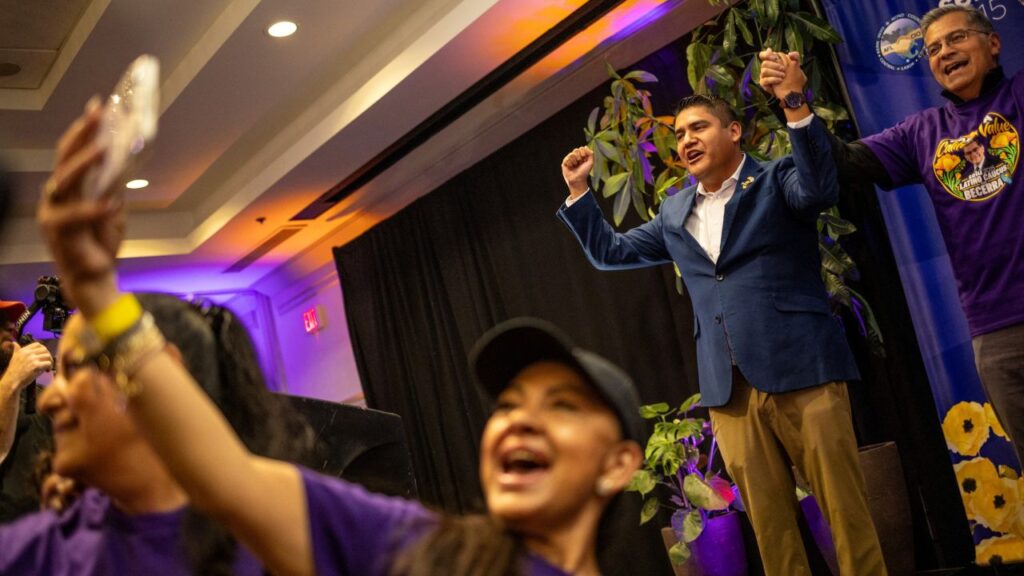 Image of two men standing on a stage with their arms raised and locked while people below take selfie pictures