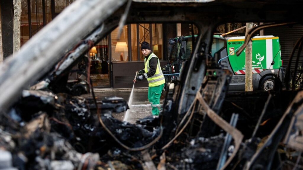 A municipal worker, seen through a burnt-out car, cleans a street a day after clashes during a national day of demonstrations against the cost of living and rising fuel prices in Paris, France, October 18, 2026. REUTERS/Benoit Tessier