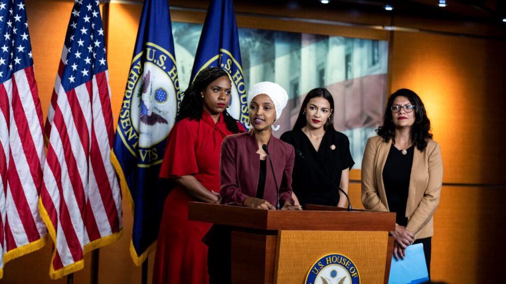 Image of four women standing on stage at a microphone with U.S. and House of Representatives flags behind them