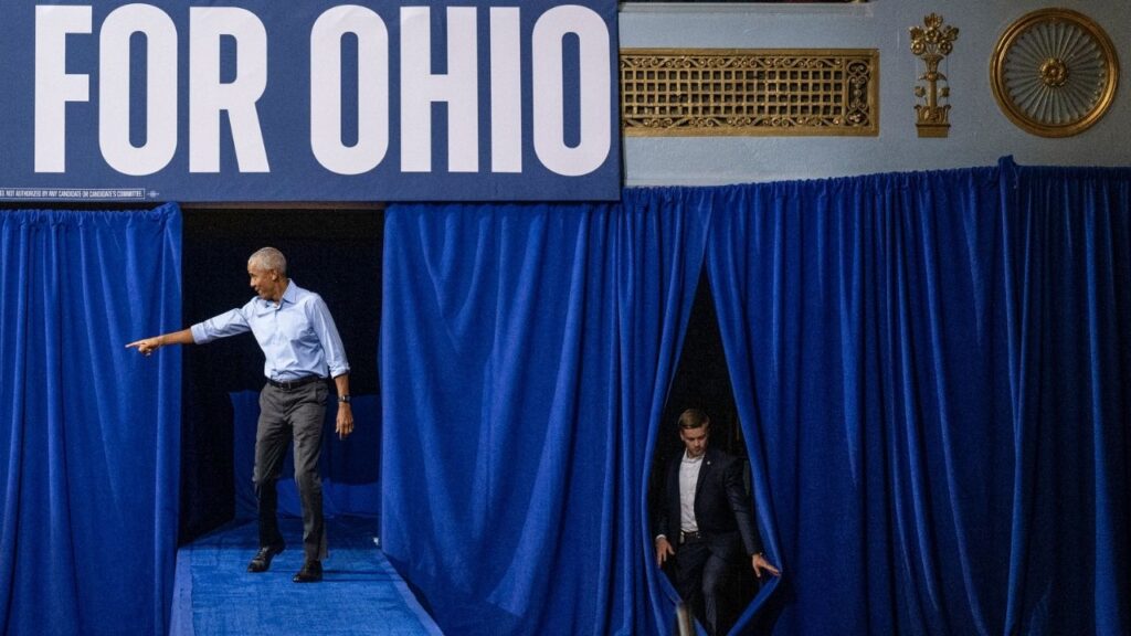 Former President Barack Obama, left, arrives on stage at a campaign rally as a member of his security detail uses a gap in the curtains in Cleveland, Ohio, Oct. 24, 2026. Eight days before the midterm elections, the jitters have taken root. (Victor J. Blue/The New York Times)