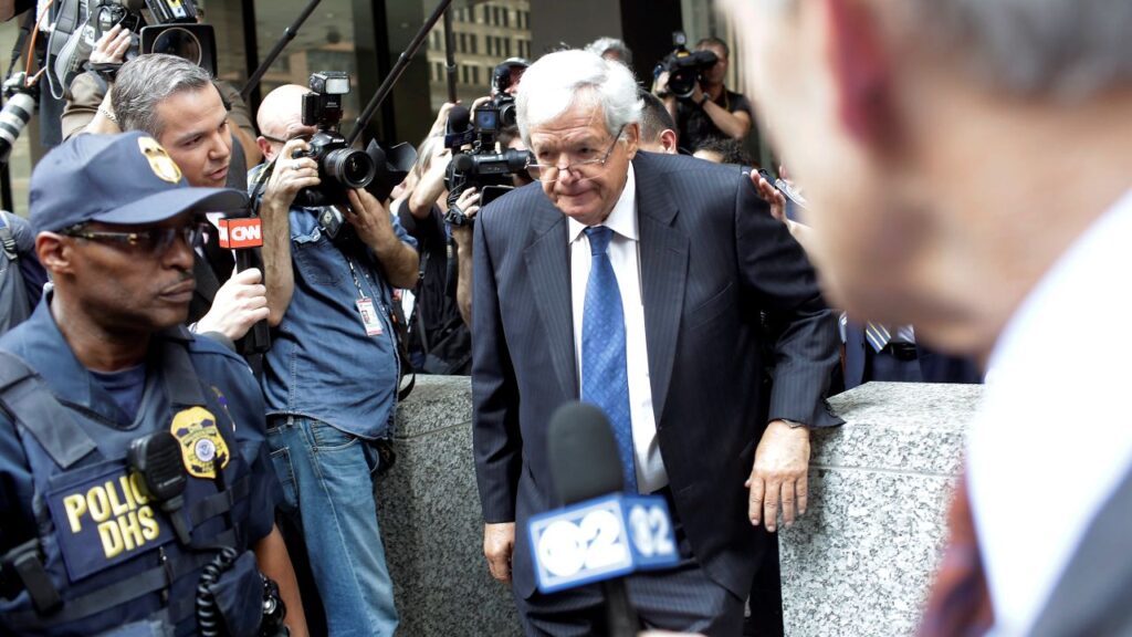 Image of Dennis Hastert in a gray suit walking out of courthouse surrounded by media and police
