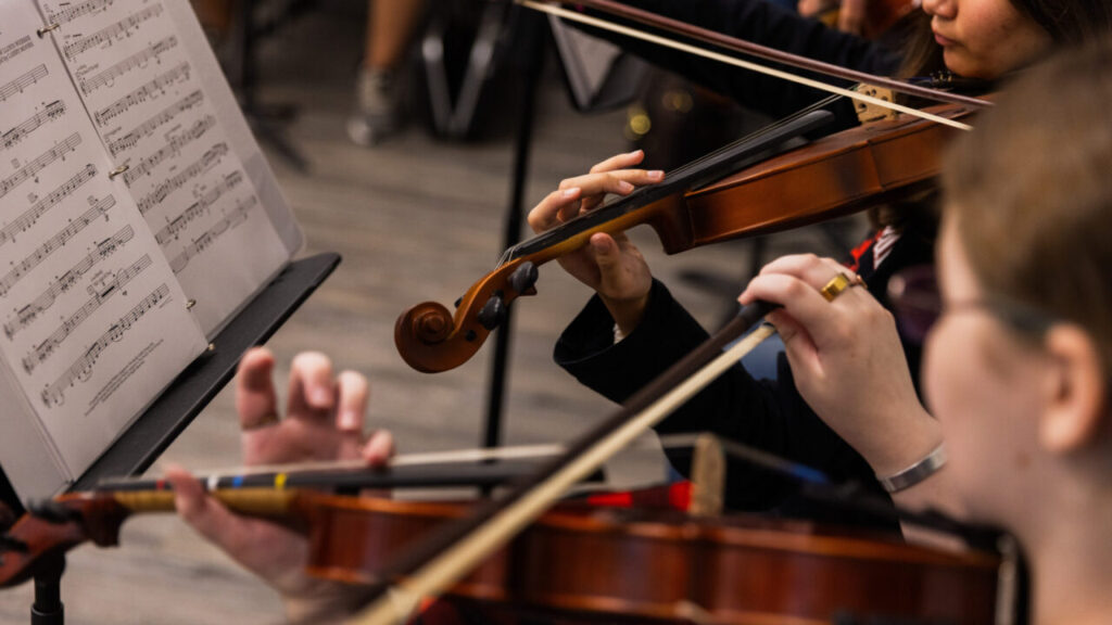 Image of high school students playing violins with sheet music placed on stands in front of them