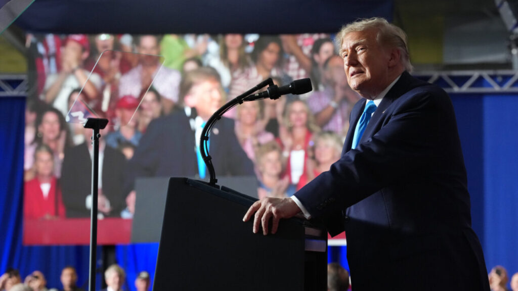 Image of President Trump in a dark suit facing left and speaking to a rally in front of a video screen livestreaming the event