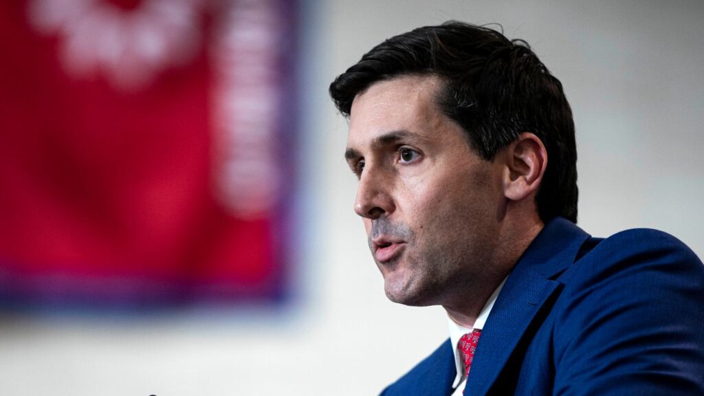 Image of a man in a blue suit facing left and sitting behind a microphone