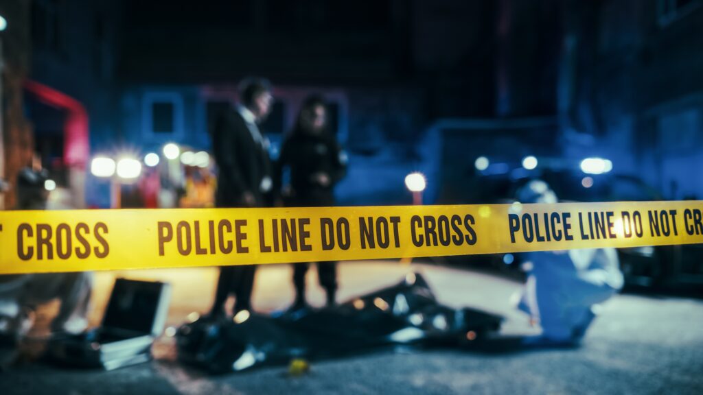 Image of yellow Police Line Do Not Cross Yellow Crime Scene tape set up at night with lights and detectives in the background